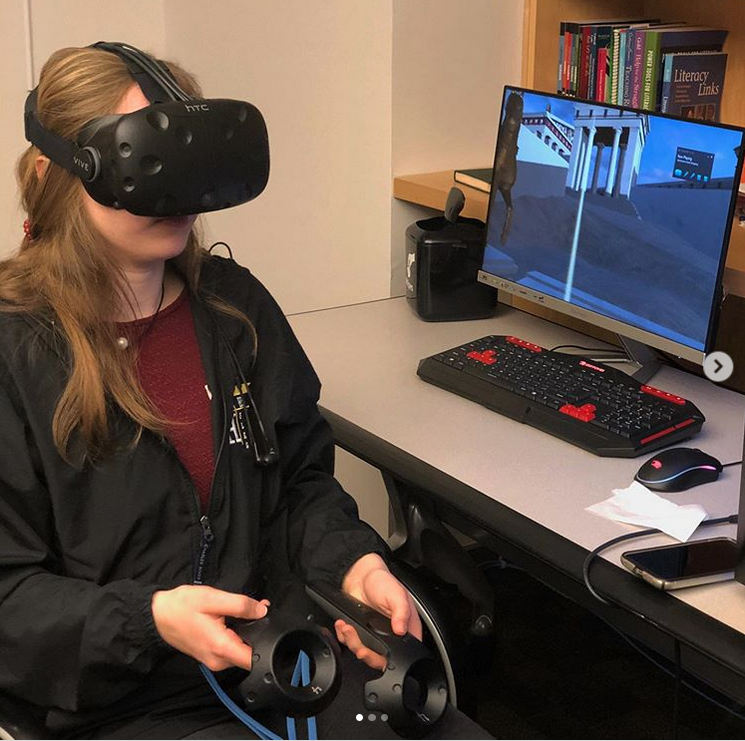 A student sitting at a VR station in the VR classroom. The student wears a headset and holds a controller in each hand. A computer monitor shows the VR program being used is an exploration of a historical site.