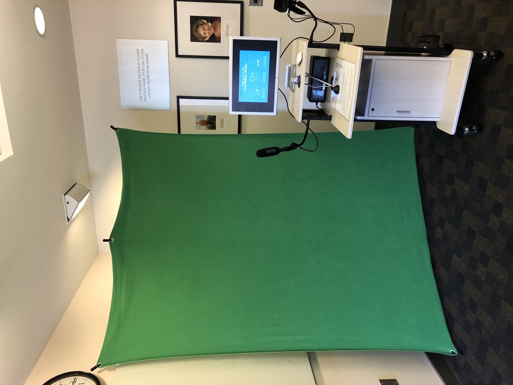 The One Button Studio, a computer monitor, camcorder, and microphone mounted on a rolling cart, sitting in front of a greenscreen.