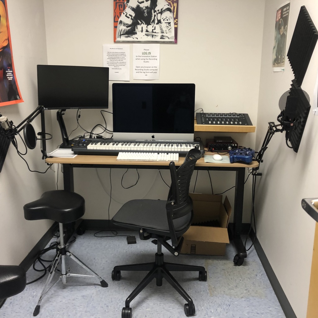 The recording studio, a room with two computer monitors, a midi keyboard, a preamp device, two seats, and two microphones.