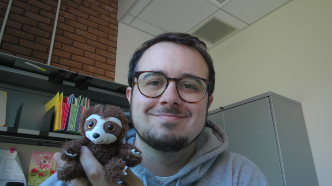 A photo of the Discovery and Electronic Resources Librarian, smiling and holding a small plush sloth in his office.