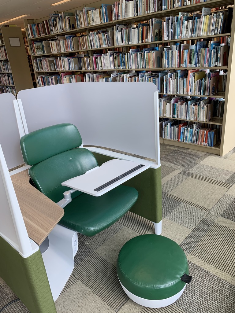 A cubicle chair consisting of one green padded seat, a rotating desk surface, a foot rest, and privacy screens built in around the chair