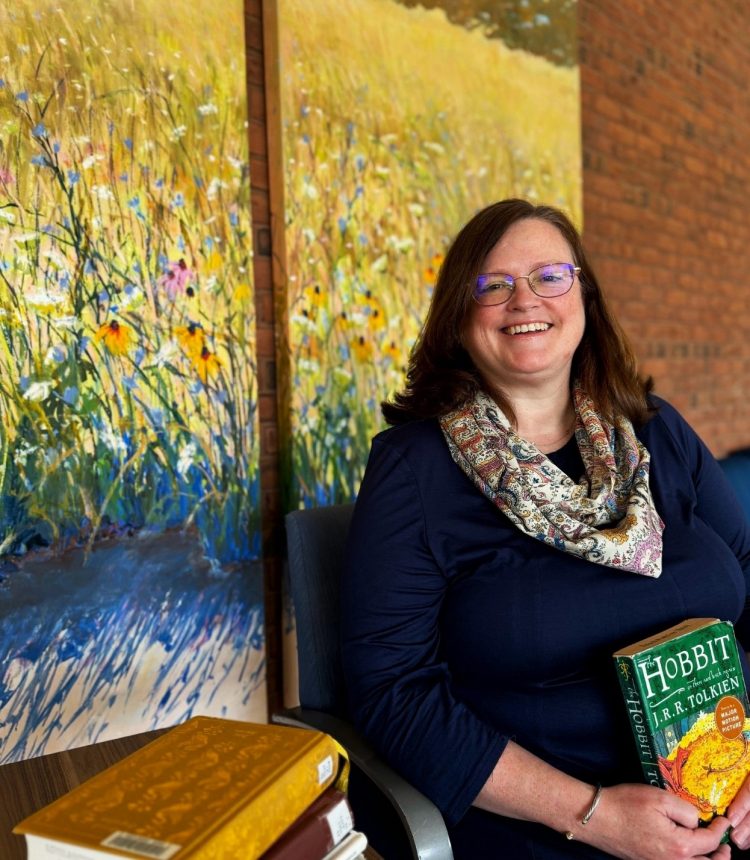 Library director Katy O'Neill sitting in a char in front of a brick wall holding a copy of The Hobbit. A pile of other books sits on a table beside her.