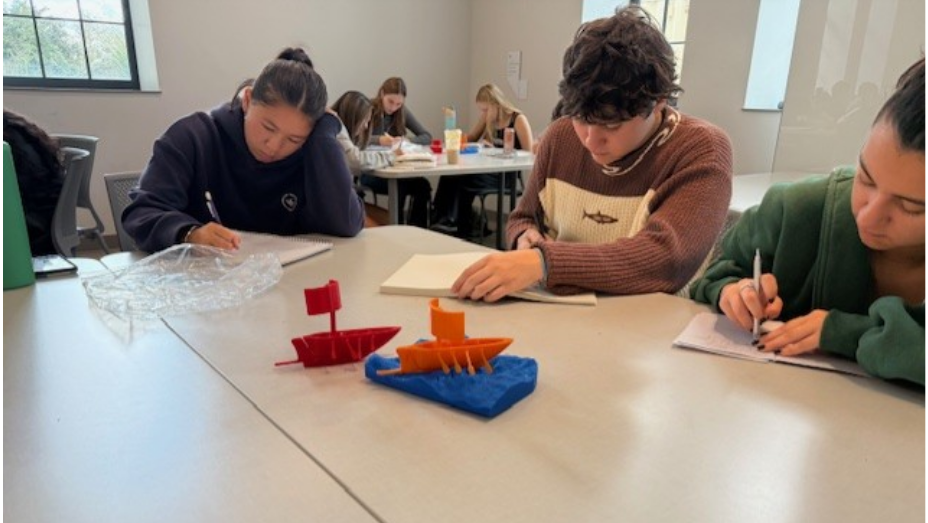 Students working around a table. In the center of the table are two different colored 3D printed ships