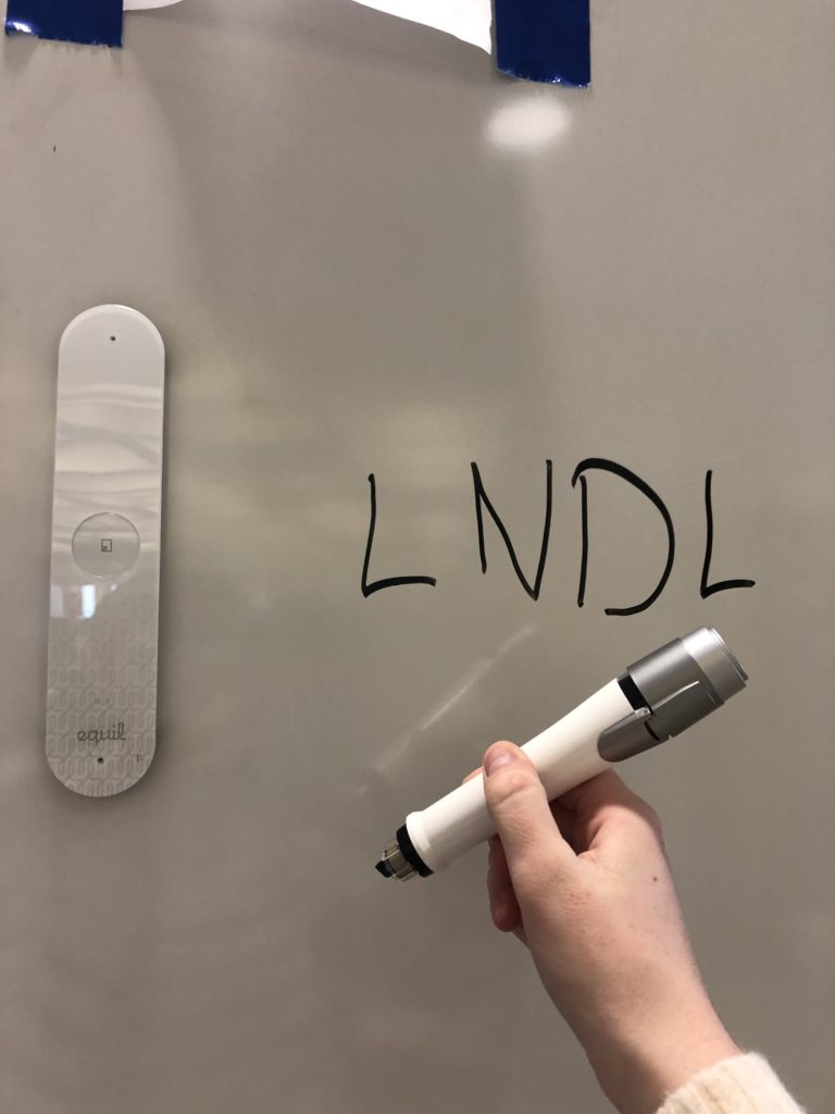 The Smartmarker's magnetic sensor is affixed to a whiteboard. A hand holds the Smartmarker in front of the whiteboard. 'LNDL' is written on the whiteboard next to the sensor.