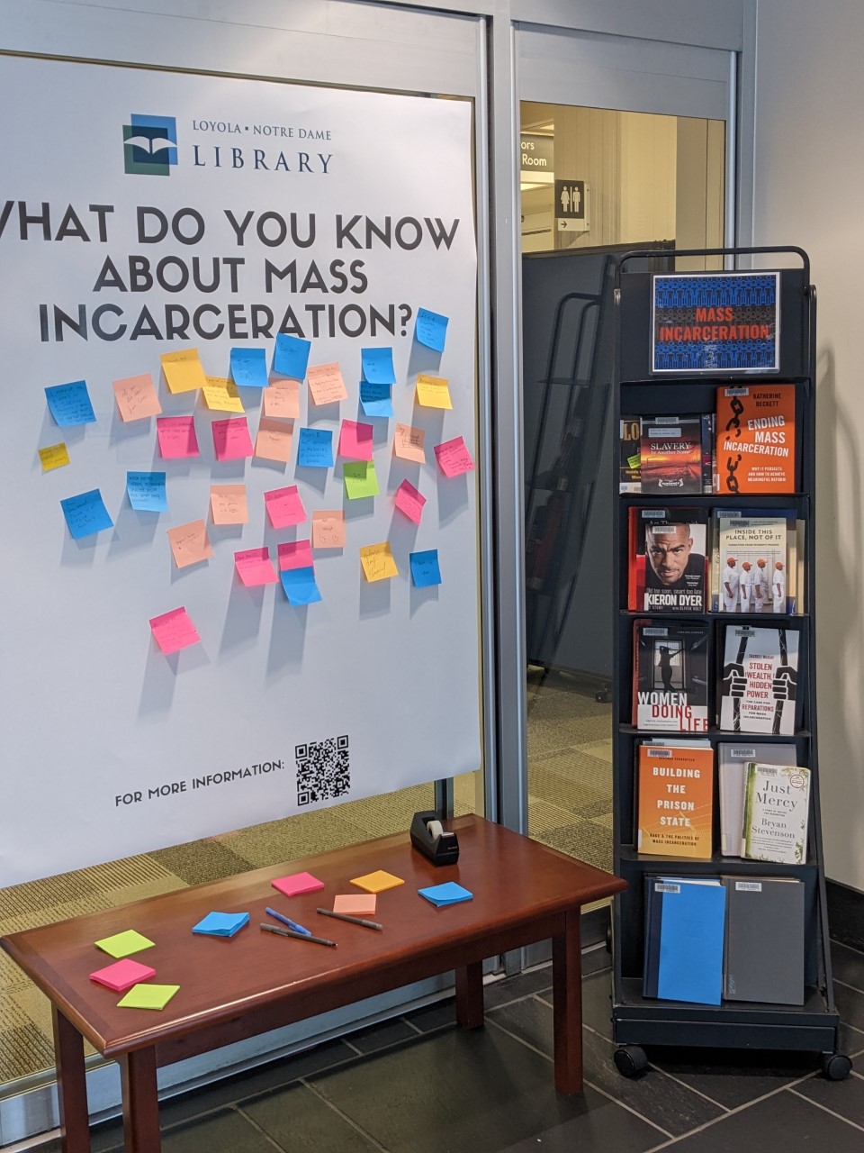 The library's exhibit and book display on the topic of mass incarceration. A large poster on the left reads 'What do you know about mass incarceration?' About two dozen post it notes filled out by patrons are stuck to the poster. To the right of the poster, a shelf displays a variety of books on the topic of mass incarceration. 